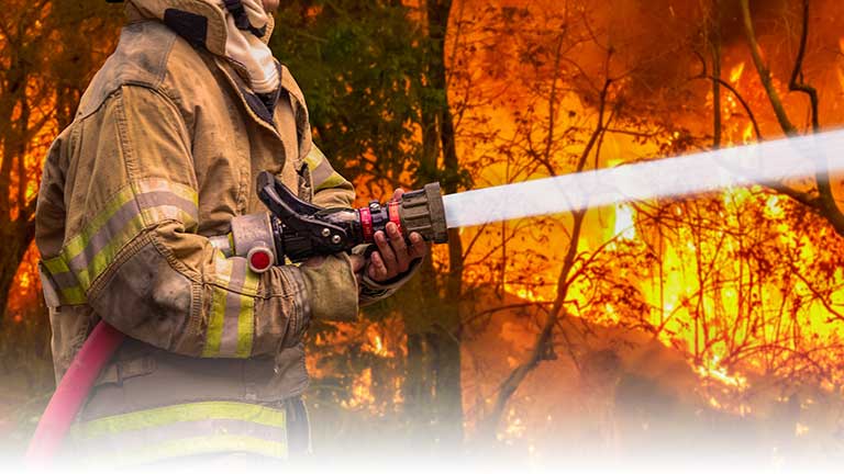 A wildfire firefighter spraying flames with water from a large hose.