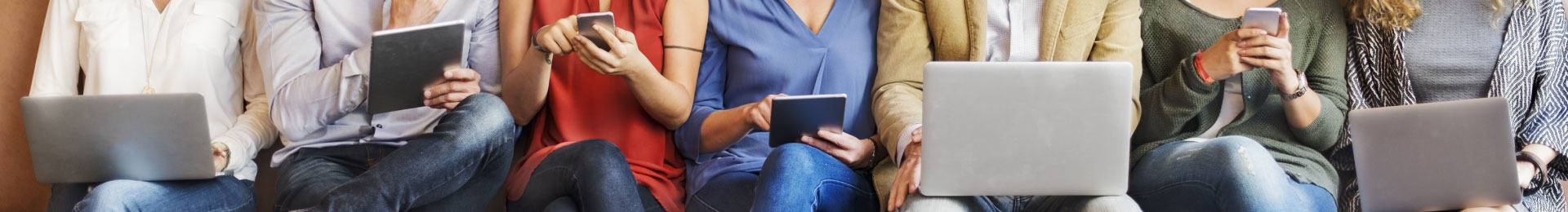 Several people seated on a bench using phones, tablets, and laptops.